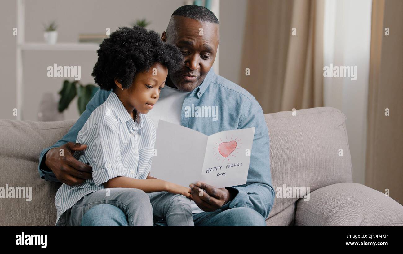 L'uomo afroamericano maturo con la ragazza carina del capretto che abbraccia seduta nella stanza sul padre del sofà legge la scheda di saluto che ama la figlia fatta il regalo con le proprie mani per Foto Stock