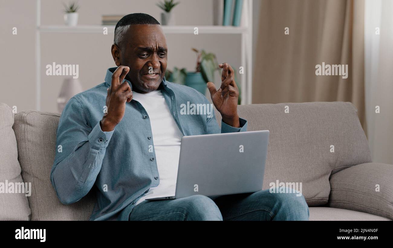 Uomo afroamericano maturo seduto a casa guardando lo schermo del laptop con speranza barrette incrociate che pregano per buona fortuna spera successo vittoria Foto Stock
