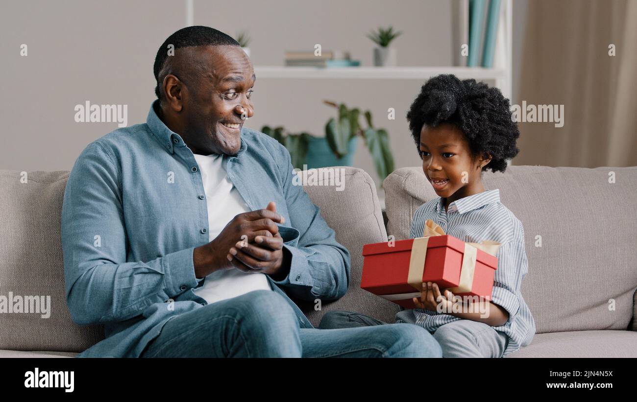 Piccola figlia felice seduta sul divano con gli occhi chiusi che riceve la scatola del regalo dal padre amorevole che cura il papà che si congratula con la ragazza del capretto che fa il compleanno Foto Stock