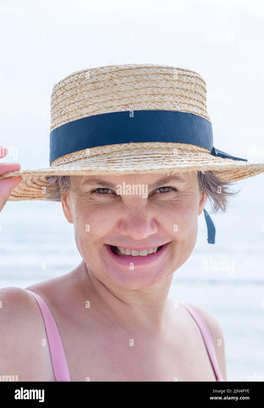 Ritratto di felice donna dai capelli grigi che sorregge con la mano il bordo del suo boater dalla brezza marina, sorridendo alla macchina fotografica. Concetto di felicità. Foto Stock