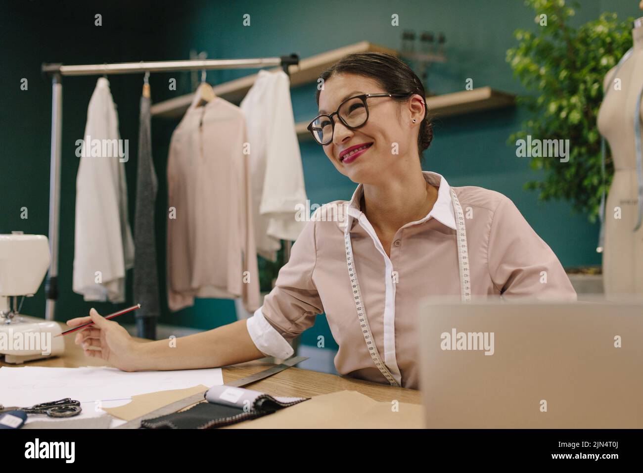 Stilista di abbigliamento seduto alla sua scrivania in studio di moda e guardando via. Donna dressmaker seduta alla sua scrivania. Foto Stock