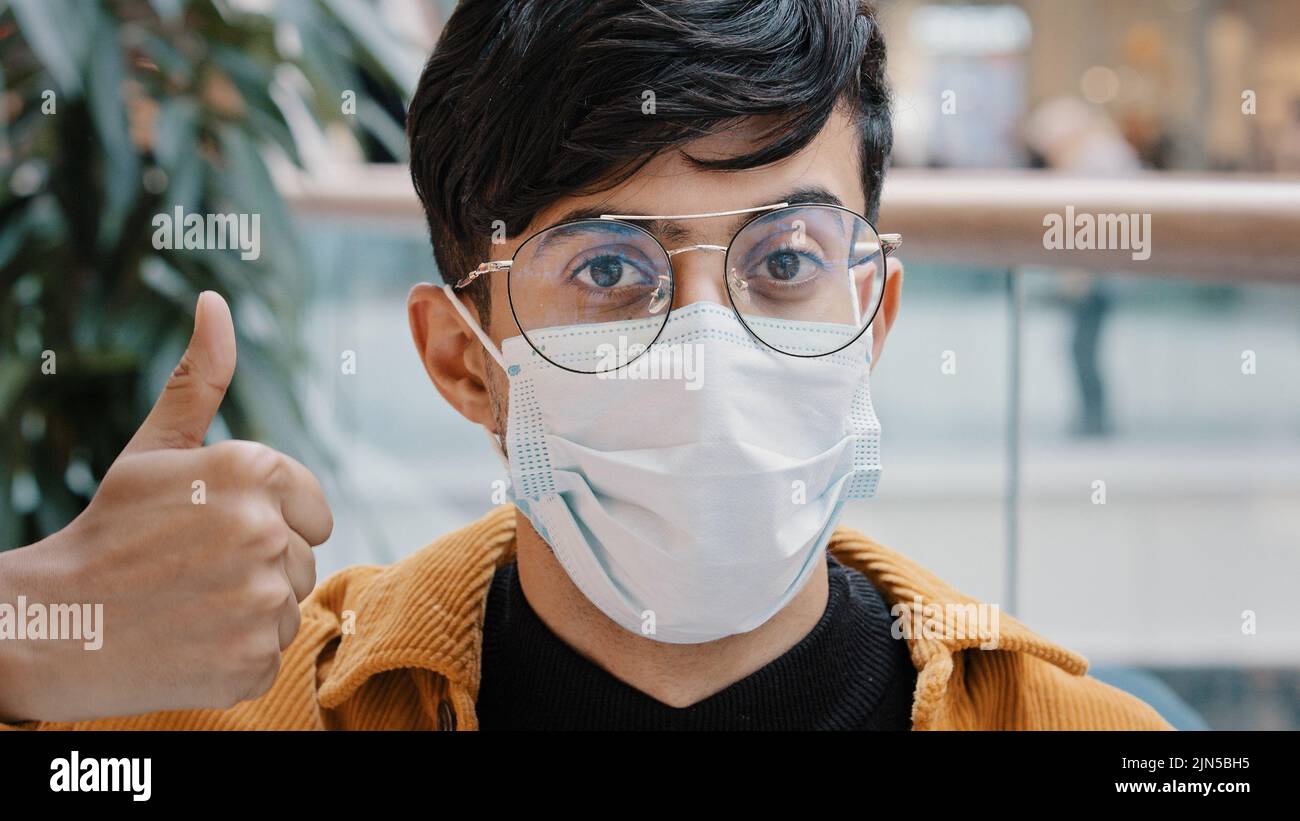 Primo piano ritratto giovane uomo in piedi in casa guardando la macchina fotografica posa indossare maschera medica monouso mostrando i pollici in su dimostra il gesto di Foto Stock