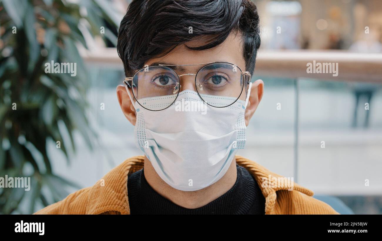 Primo piano ritratto giovane uomo in piedi al chiuso guardando la macchina fotografica posa indossare maschera medica monouso a causa di covid19 virus pandemic focolaio consapevole Foto Stock