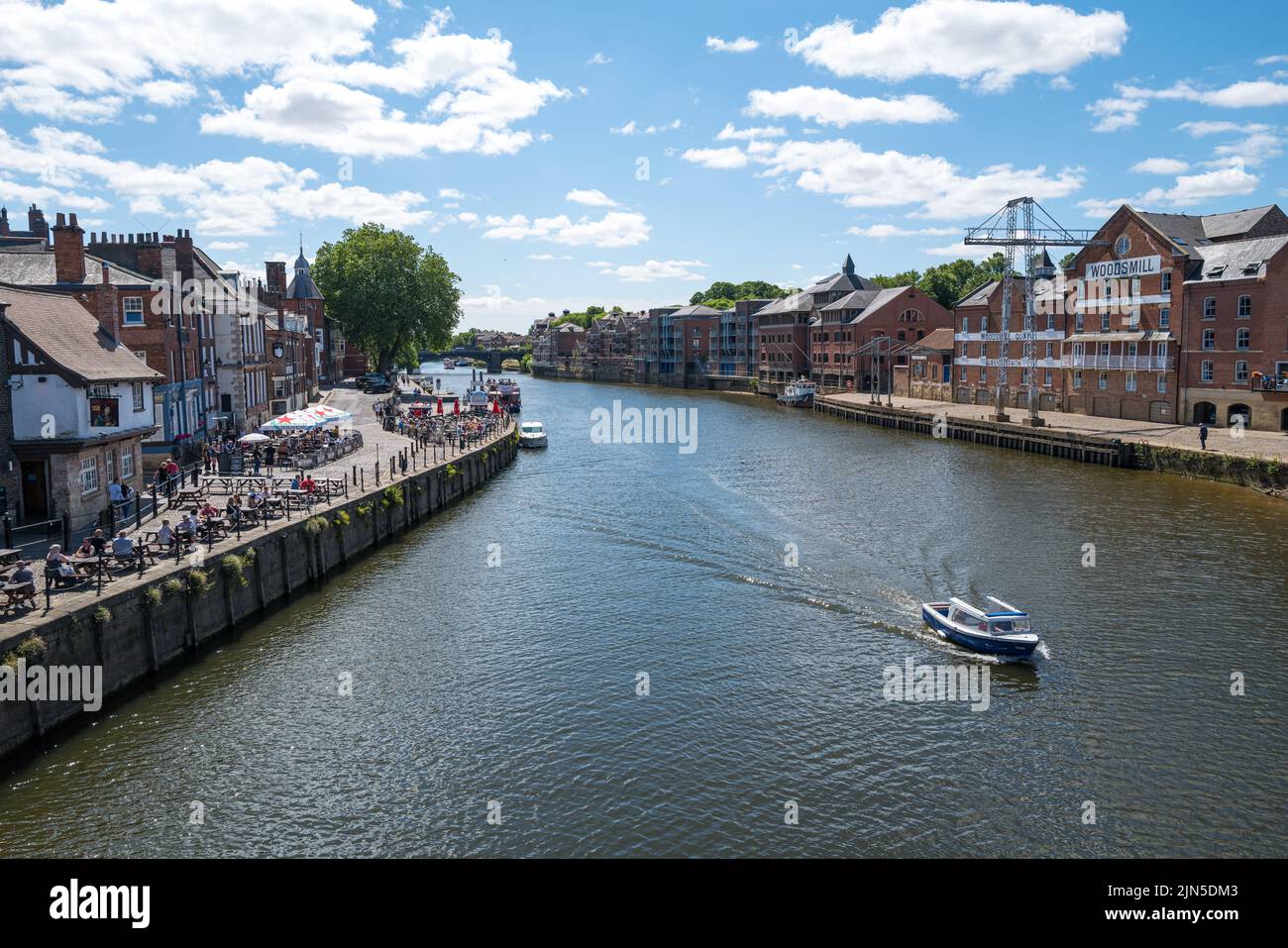 Una barca sul fiume passa accanto al pub Kings Arms, dove i clienti si siedono all'esterno e bevono un drink nel centro di York, di fronte ai vecchi magazzini di grano Foto Stock