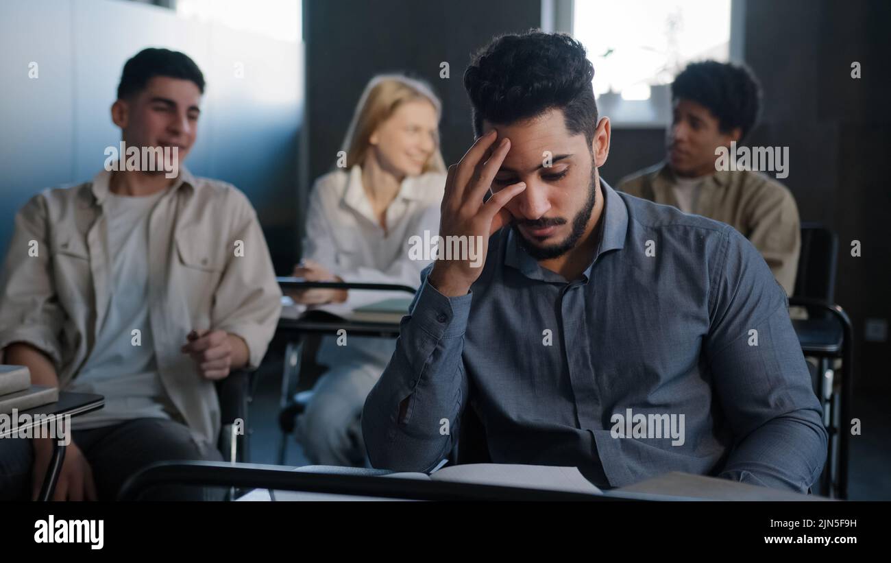 Triste frustrato studente arabo seduto in classe alla scrivania solo che soffre di maltrattamenti da parte dei compagni di classe discriminazione razziale si sente vittima di bullismo Foto Stock