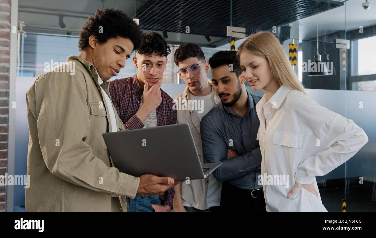 Il giovane dipendente dell'azienda spiega la strategia di sviluppo racconta nuove idee mostrando il piano di lavoro sul notebook a un team creativo sorridente e diversificato Foto Stock