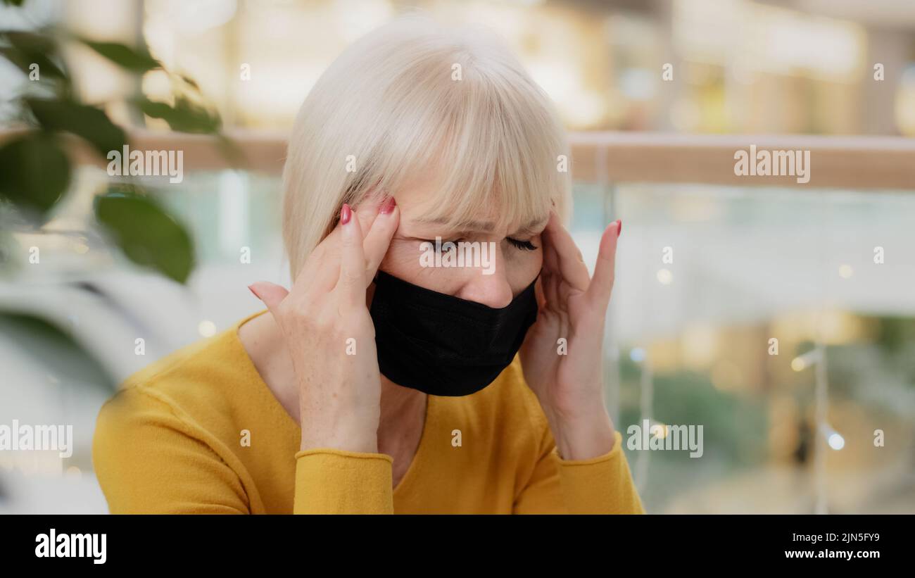 Primo piano infelice donna caucasica di mezza età in maschera medica seduta indoor tenendo le mani dietro la testa soffrendo mal di testa chiusura occhi sensazione di stanchezza Foto Stock