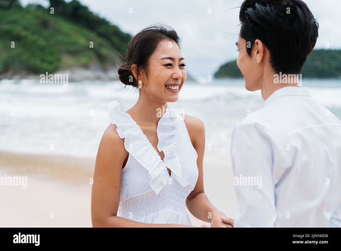 Felice toothy sorridente asiatico 30s coppia in rapporto in candid sulla spiaggia in estate. Una coppia romantica che indossa una camicia bianca e un vestito. Foto Stock