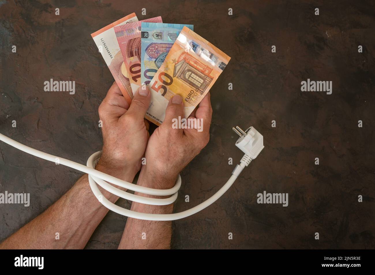 Mani di un uomo con banconote in euro e sono legati con un cavo di alimentazione elettrica, concetto per l'aumento dei costi dell'elettricità e risparmio energetico, backgrou scuro Foto Stock