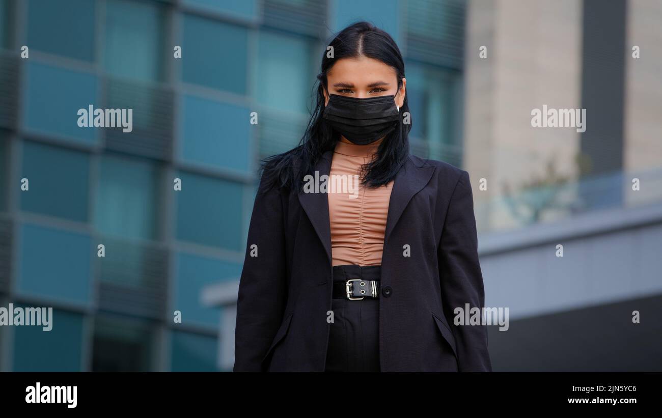 Giovane ragazza ispanica in piedi sulla città edificio sfondo guardando fotocamera businesswoman indossare maschera medica protettiva protegge la salute dal virus Foto Stock