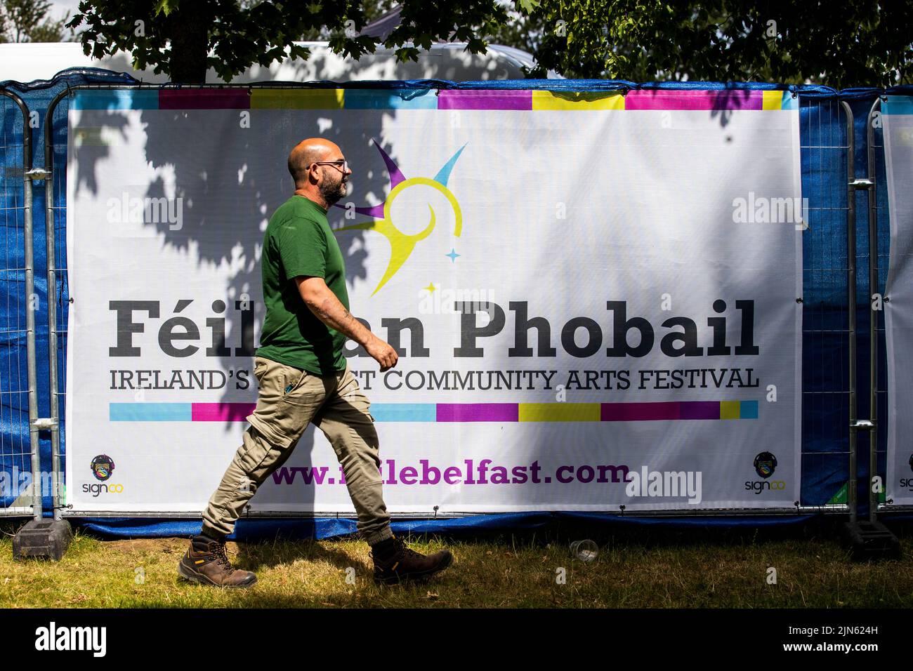 Un uomo cammina davanti a un banner per Feile An phobail, noto anche come West Belfast Festival, nel Falls Park. Foto Stock