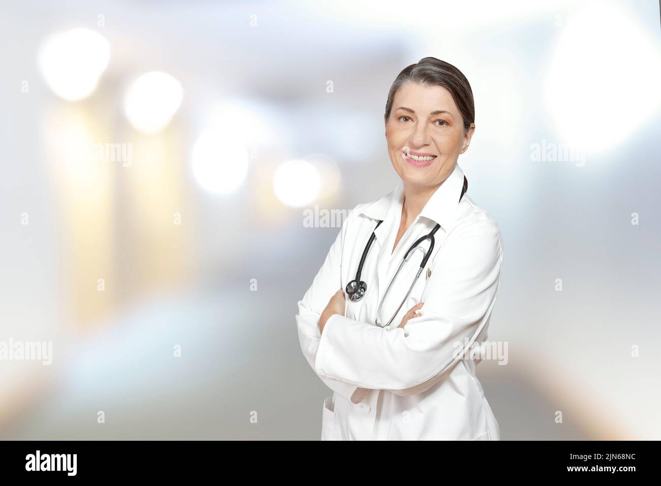 Medico matura in un camice da laboratorio bianco, sorridente amichevole, contro lo sfondo sfocato di un centro medico interno. Foto Stock