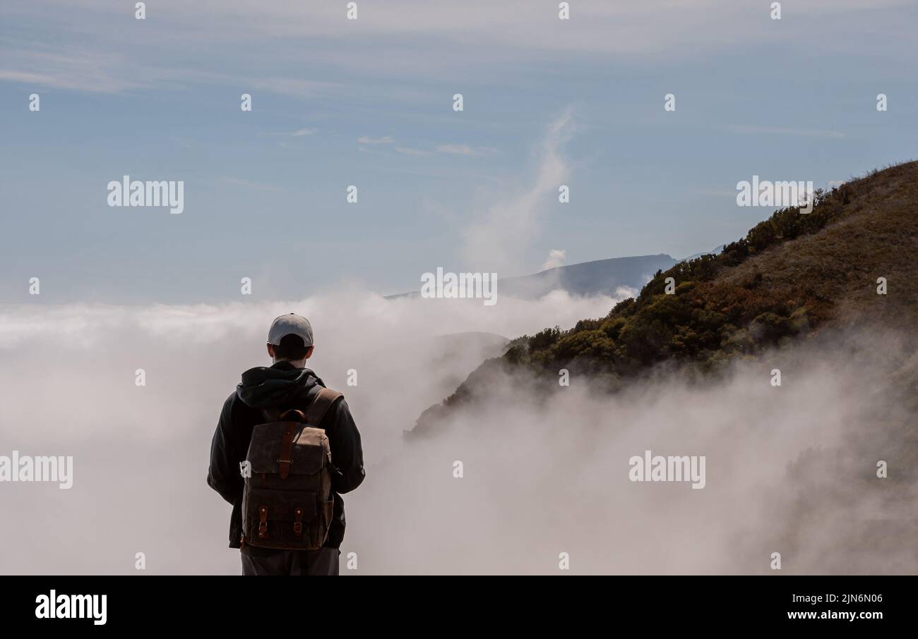 Giovane uomo con uno zaino turistico che guarda un paesaggio panoramico di montagne e nuvole, escursioni a Madeira Foto Stock