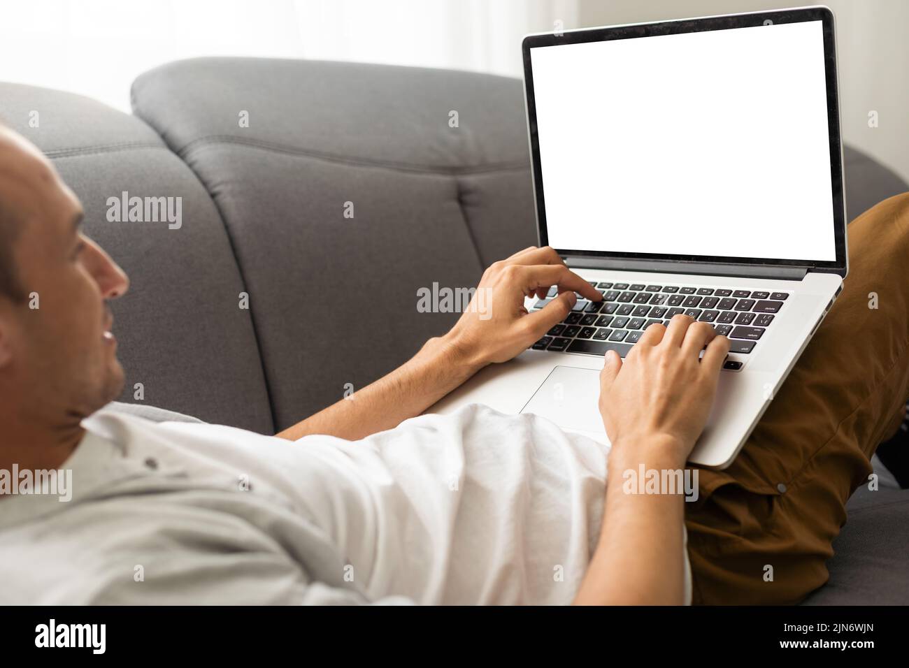 Giovane utente tecnico maschile che si rilassa sul divano che tiene un computer portatile mock up bianco schermo bianco. Uomo che usa il moderno notebook che naviga in internet, leggi le notizie Foto Stock