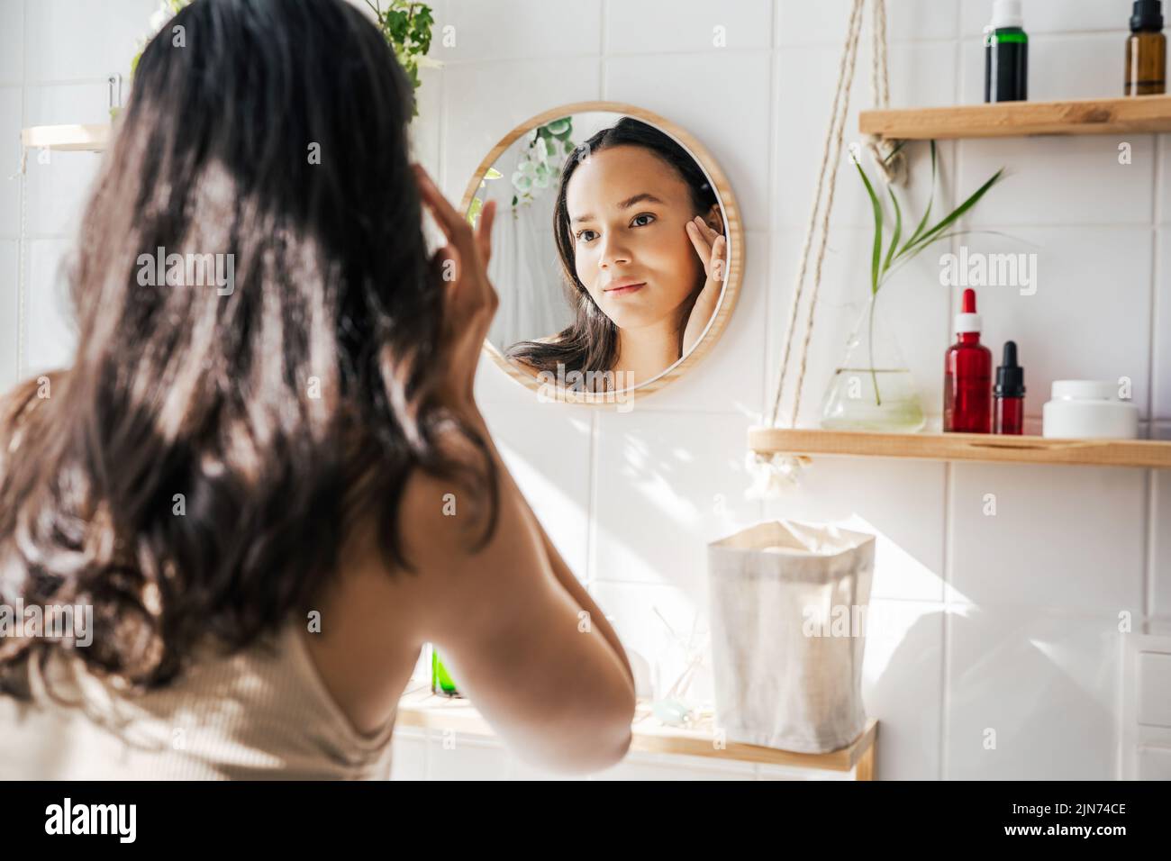 Bella donna ispanica che guarda nello specchio in bianco eco-friendly bagno. Stalle in legno e bottiglie riutilizzabili per cosmetici. Benessere, concetto di cura della pelle Foto Stock