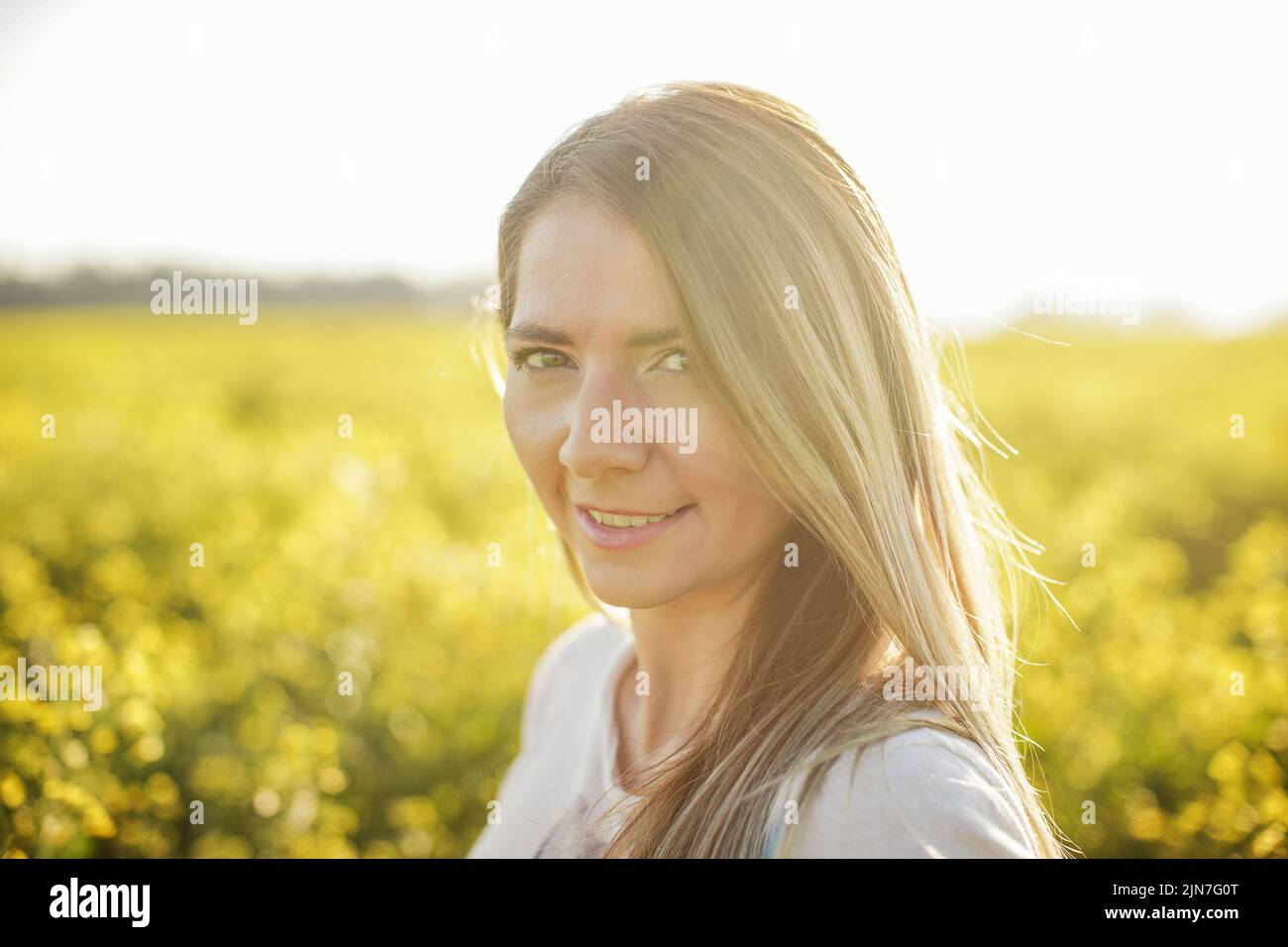 Ritratto di giovane donna con capelli lunghi, campo di fiori gialli e luce solare dietro di lei Foto Stock