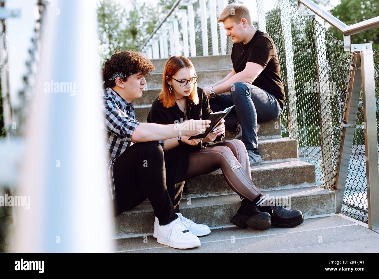 Gruppo di giovani studenti seduti su scale di cemento, preparandosi all'esame. Giovane donna, curly uomo con libro aperto. Foto Stock