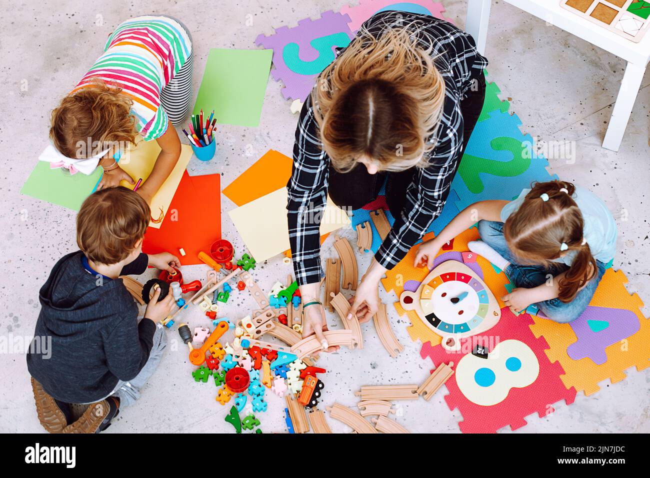 Vista dall'alto di una giovane insegnante seduta sul pavimento con i bambini degli alunni, che mostra come unirsi alla ferrovia in legno in aula. Foto Stock