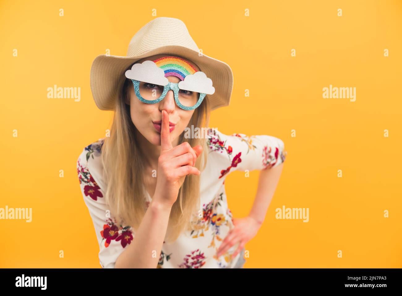 Creativa bella bionda caucasica signora con divertente arcobaleno occhiali che mostrano un segno di silenzio con il suo dito e guardando la fotocamera - sfondo arancione. Foto di alta qualità Foto Stock
