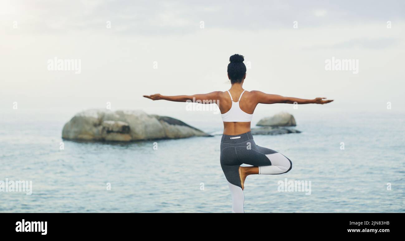 Fidarsi del mio equilibrio. Dietro l'inquadratura di una donna irriconoscibile in piedi e facendo yoga da sola sull'oceano durante una giornata trascorsa. Foto Stock