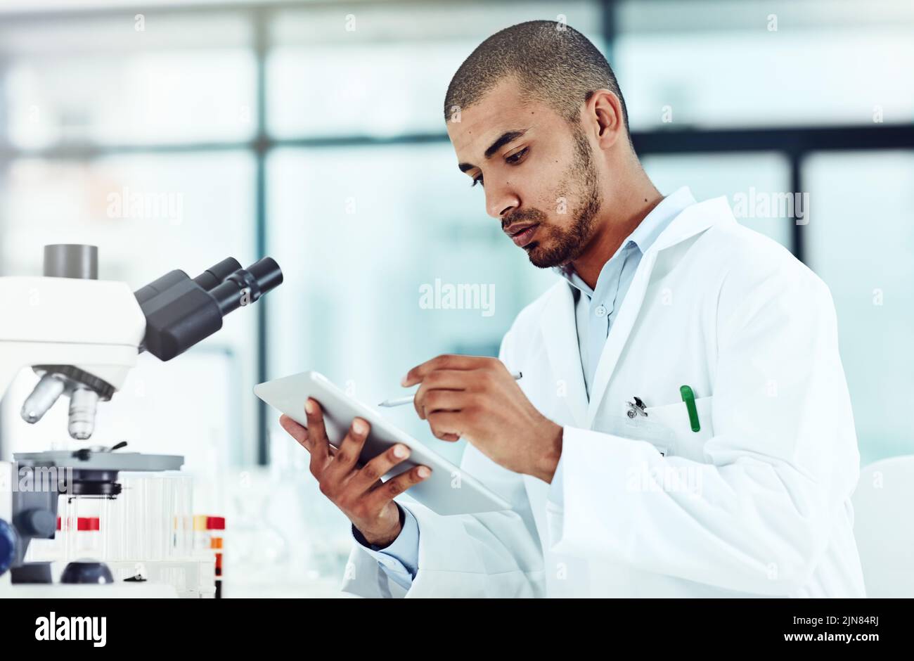 Serio scienziato maschile che lavora su un tablet rivedendo una pubblicazione di dottorato online in un laboratorio. Lavoratore di laboratorio che aggiorna i dati sanitari per una rivista scientifica Foto Stock