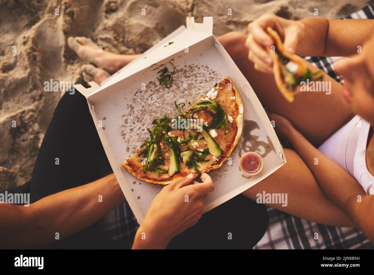 A metà strada, una giovane coppia irriconoscibile che ha una piccola data di picnic e che condivide una pizza sulla spiaggia al tramonto. Foto Stock