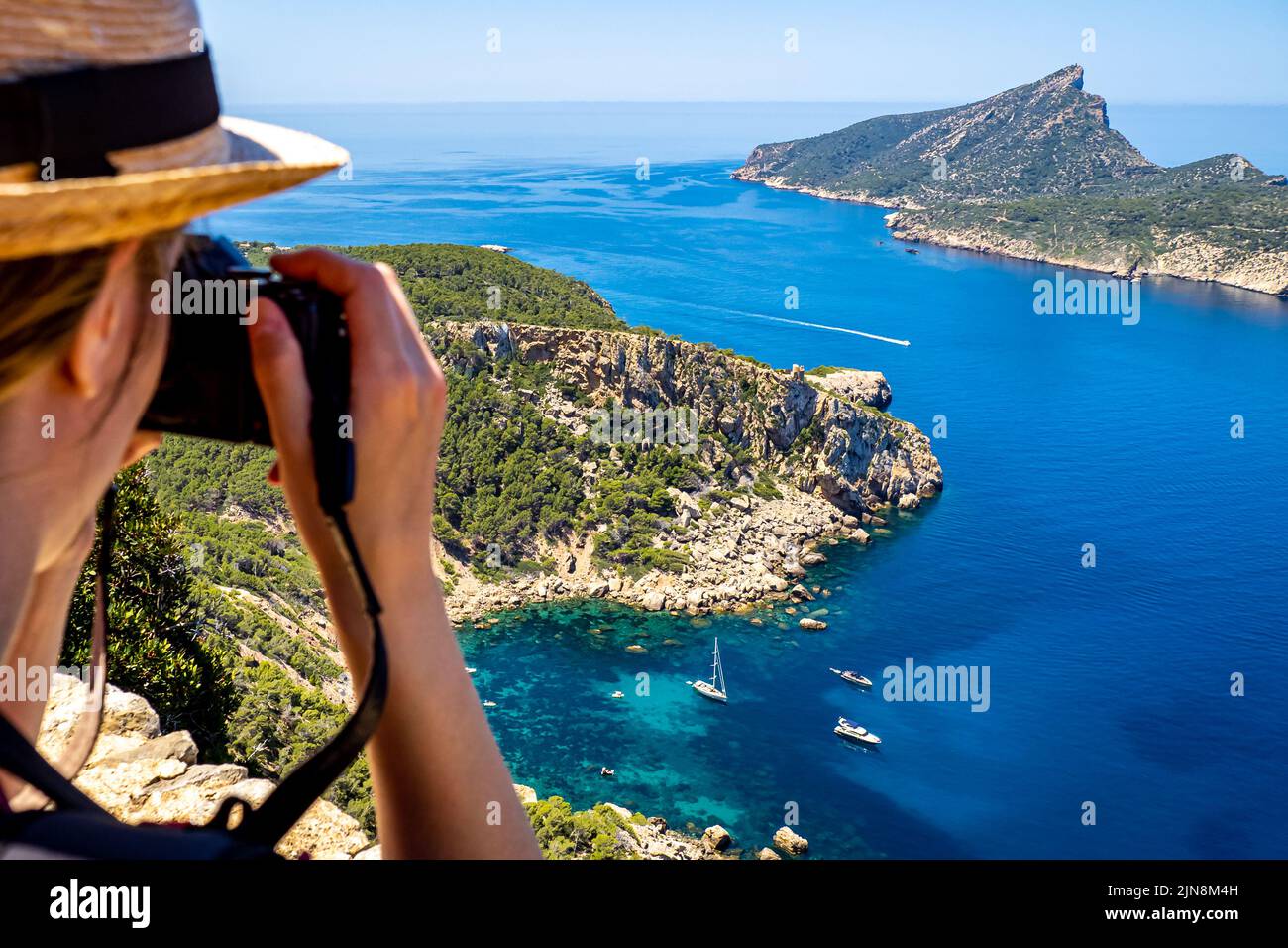 Vista posteriore di una giovane donna bianca con una macchina fotografica e un cappello sopra l'accogliente baia Cala en Basset vicino al villaggio di Sant Elm, nella parte più occidentale di Maiorca Foto Stock