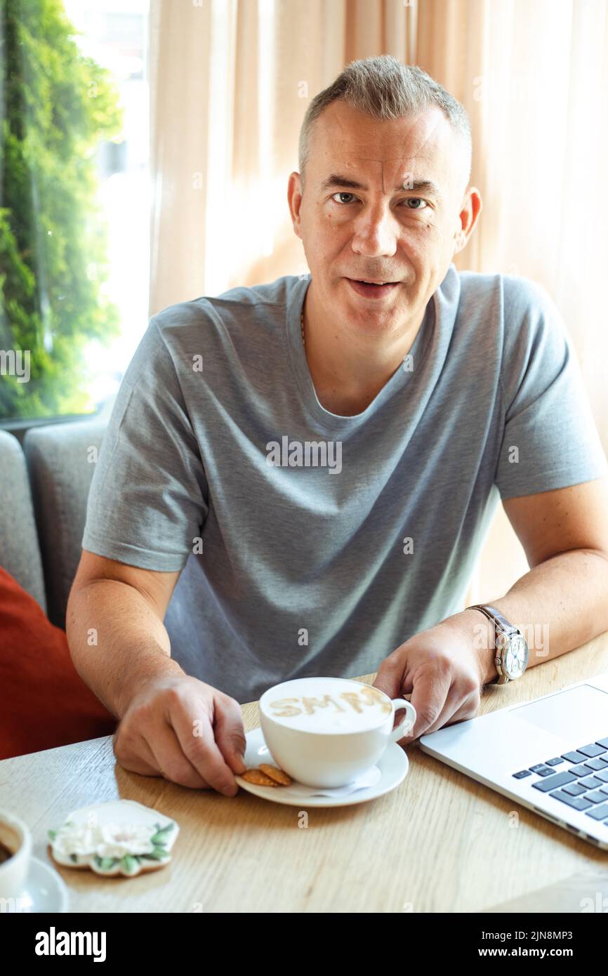 Uomo d'affari anziano e bello seduto in un bar con una tazza di caffè e lavorando su un computer portatile. Social media marketing, SMM, promozione, lavoro remoto, aromatizzato Foto Stock