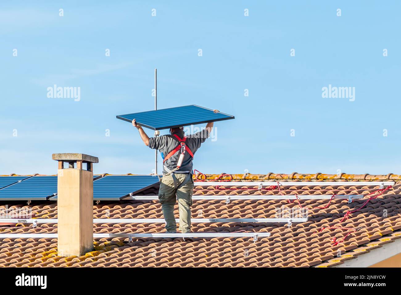 Un lavoratore solleva un pannello solare sulla sommità di un tetto con tegole rosse per installare un impianto fotovoltaico Foto Stock