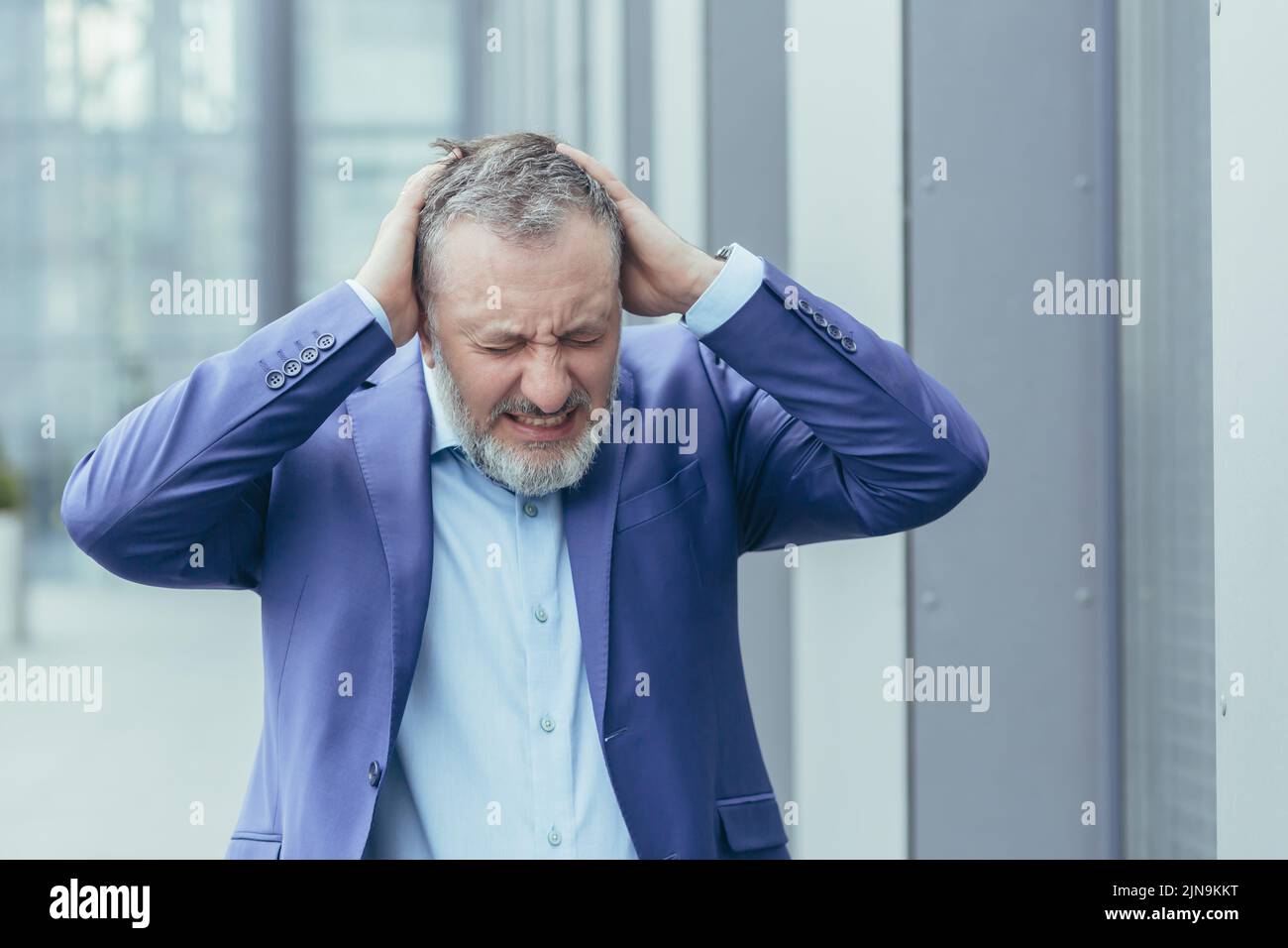 Uomo d'affari anziano dai capelli grigi fuori dall'ufficio edificio depresso, uomo in disperazione e disprezzo, investitore fallimentare perso denaro, tenendo le mani sulla testa Foto Stock