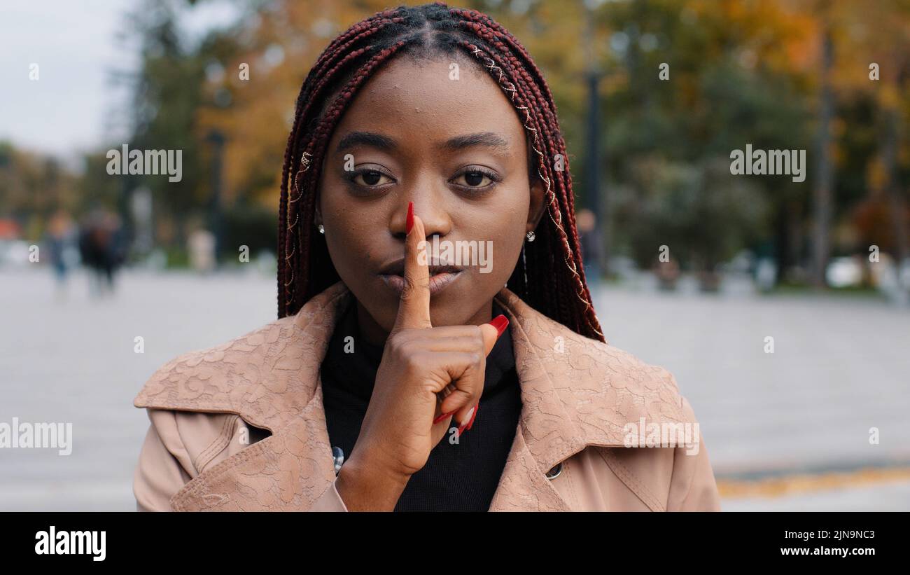 Primo piano giovane fiducioso misterioso afroamericano donna in piedi all'aperto mostrando il gesto del silenzio mette il dito contro la bocca vieta di parlare proibiti Foto Stock