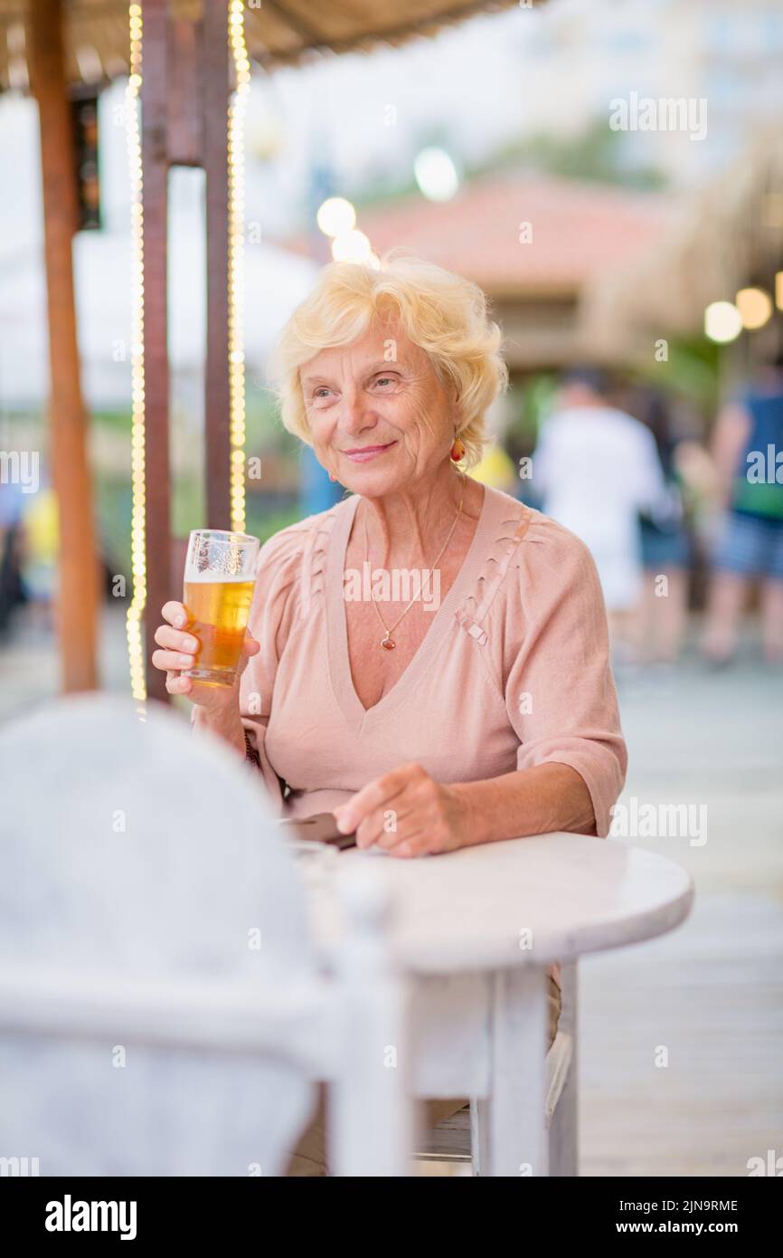 Donna matura seduta a un tavolo in un bar estivo e bere birra Foto Stock