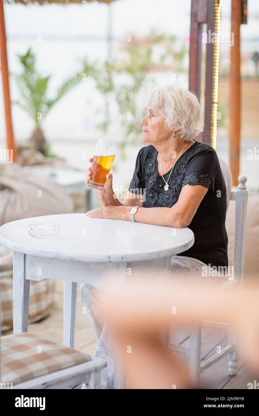 Donna matura seduta a un tavolo in un bar estivo e bere birra Foto Stock