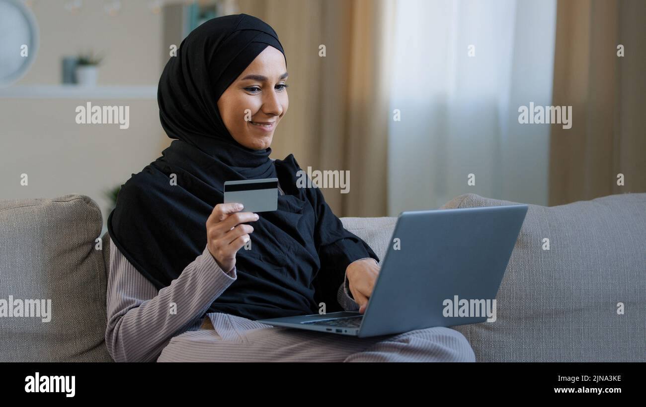 Sorridendo cliente della ragazza islamica in hijab seduta sul divano fare ordine in linea usare il laptop inserire il numero della carta di credito per la transazione felice donna musulmana pagare Foto Stock