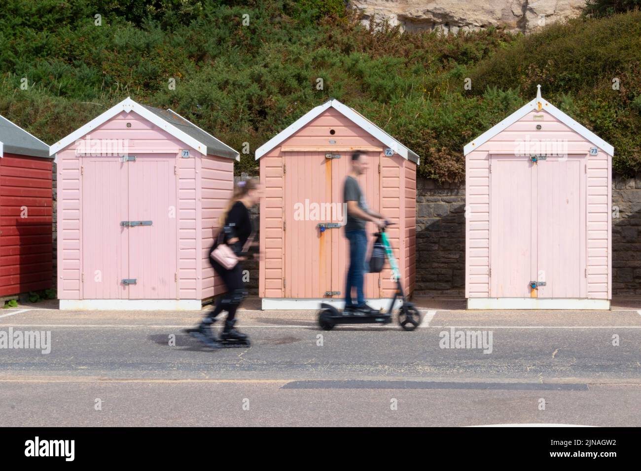 Fila di capanne in legno rosa multicolore sul mare con skater e uomo su scooter elettrico che passa di fronte, a Bournemouth Dorset UK Foto Stock