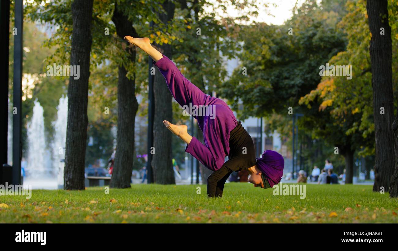 Forte attiva ragazza musulmana in hijab donna yoga sportswoman fare yoga allenamento nel parco sul prato verde facendo equilibrio handstand allenamento tenendo la gamba in aria Foto Stock