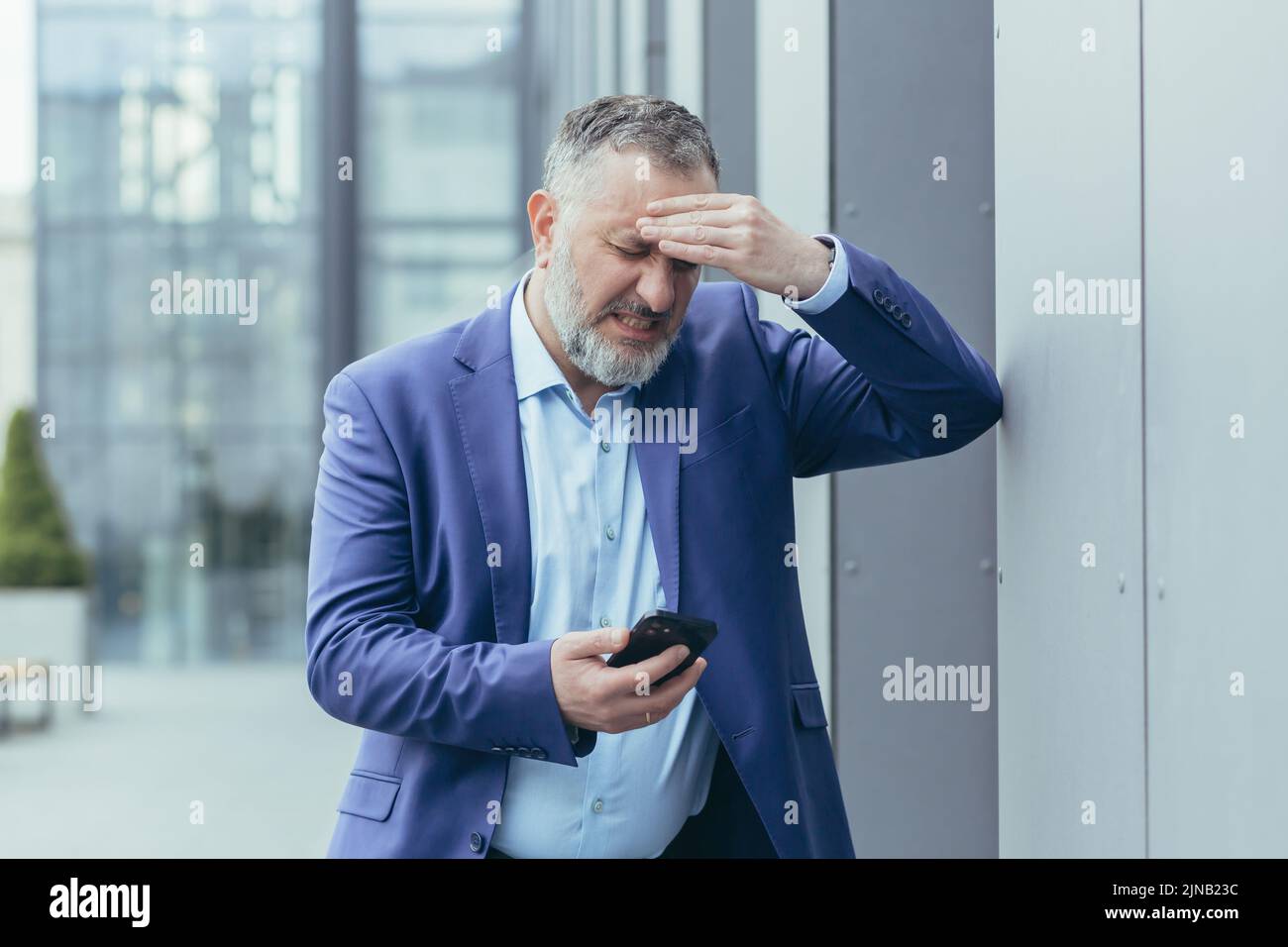 Uomo d'affari anziano grigio-haired malato fuori dell'edificio dell'ufficio, uomo che tiene la mano alla testa, avendo mal di testa grave, capo che cerca di chiamare il medico al telefono e chiamare l'ambulanza Foto Stock