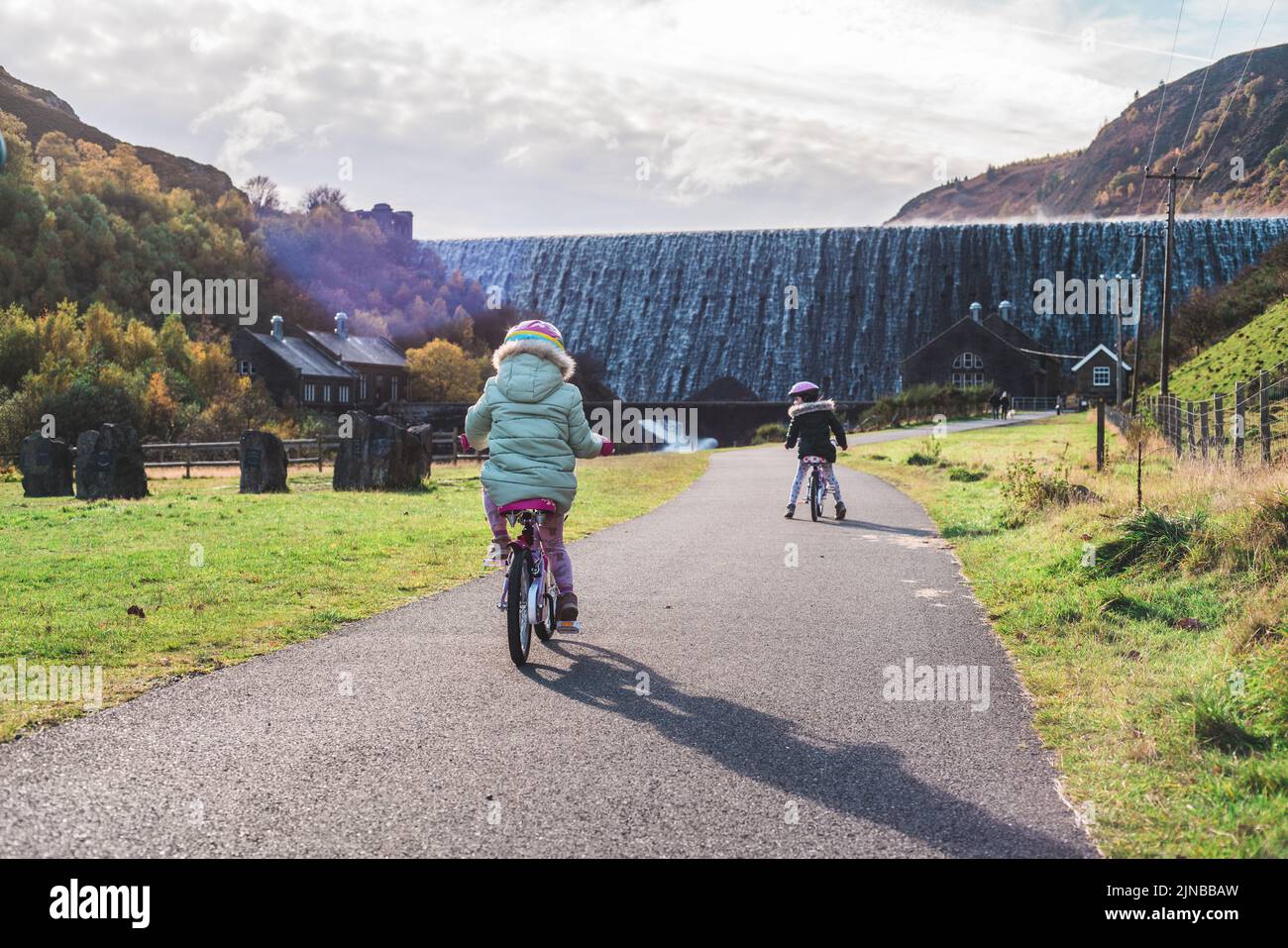 Due bambini in bicicletta con una diga sullo sfondo che ha l'acqua che scorre sopra di essa Foto Stock