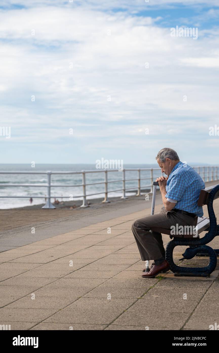 Un vecchio seduto su una panchina vicino al mare, tenendo un bastone da passeggio con gli occhi chiusi Foto Stock