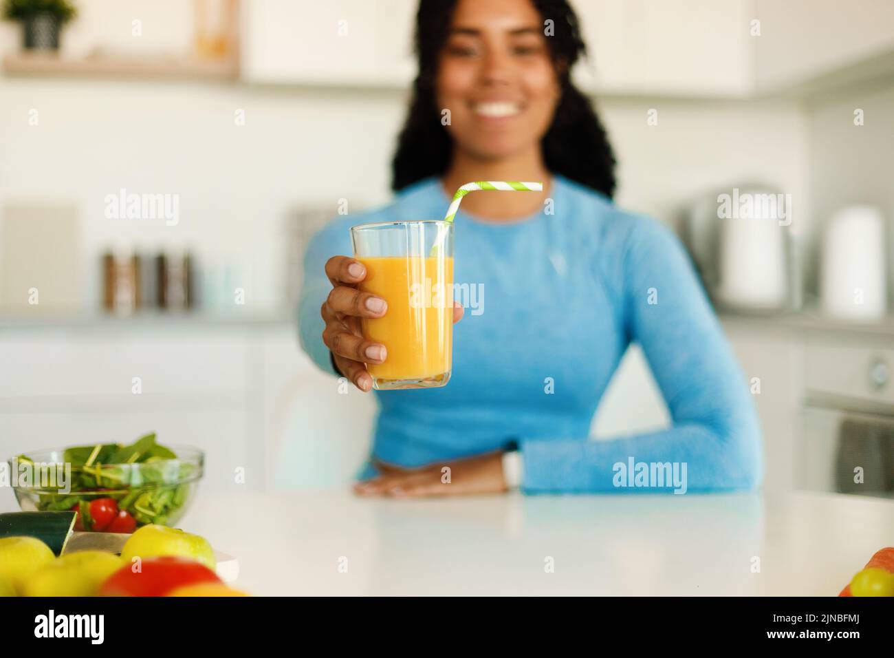 Donna felice di fitness nera in abbigliamento sportivo che mostra succo d'arancia fresco in vetro, seduta in cucina dopo l'allenamento domestico, Foto Stock