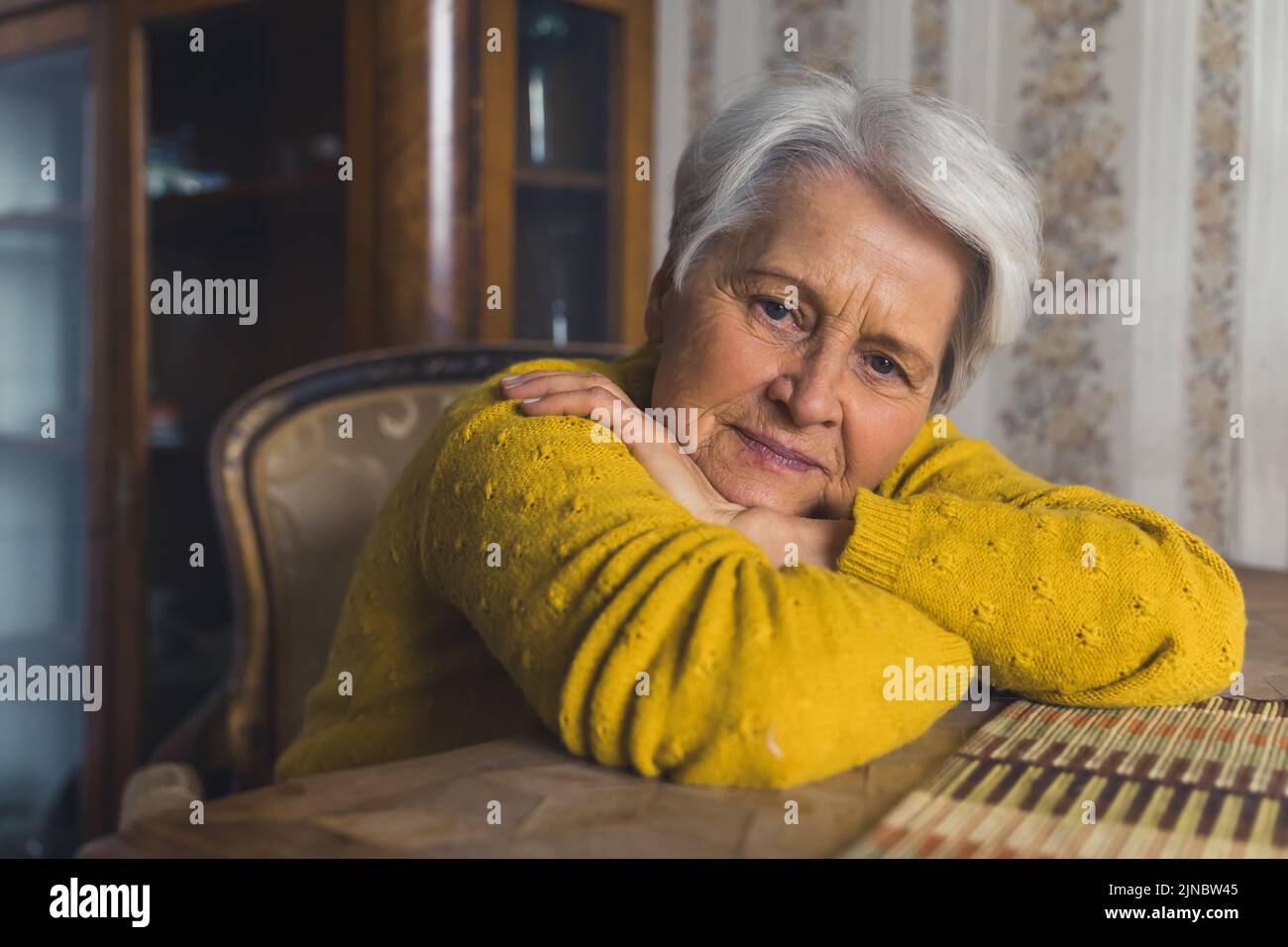 Sola vedova anziana dai capelli grigi seduta al tavolo del soggiorno con la testa appoggiata sulle braccia incrociate e pensando alla sua famiglia. Foto di alta qualità Foto Stock