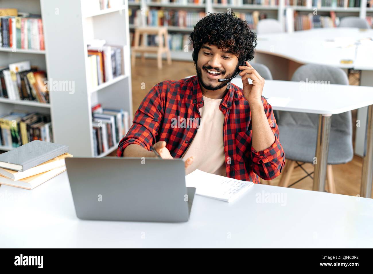 Gioioso moderno intelligente arabo o indiano studente maschio, con cuffie, si siede al tavolo con il computer portatile in biblioteca universitaria, parlando tramite videoconferenza, ascolta video corso on-line, discutere nuove informazioni Foto Stock