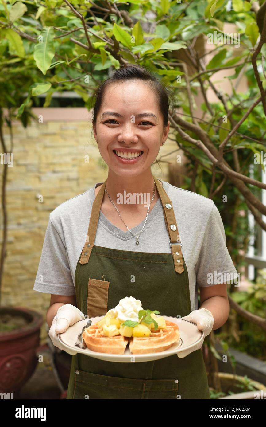 Cameriera femminile vietnamita che serve waffle belgi con frutta e gelato in una caffetteria all'aperto che guarda in alto Foto Stock