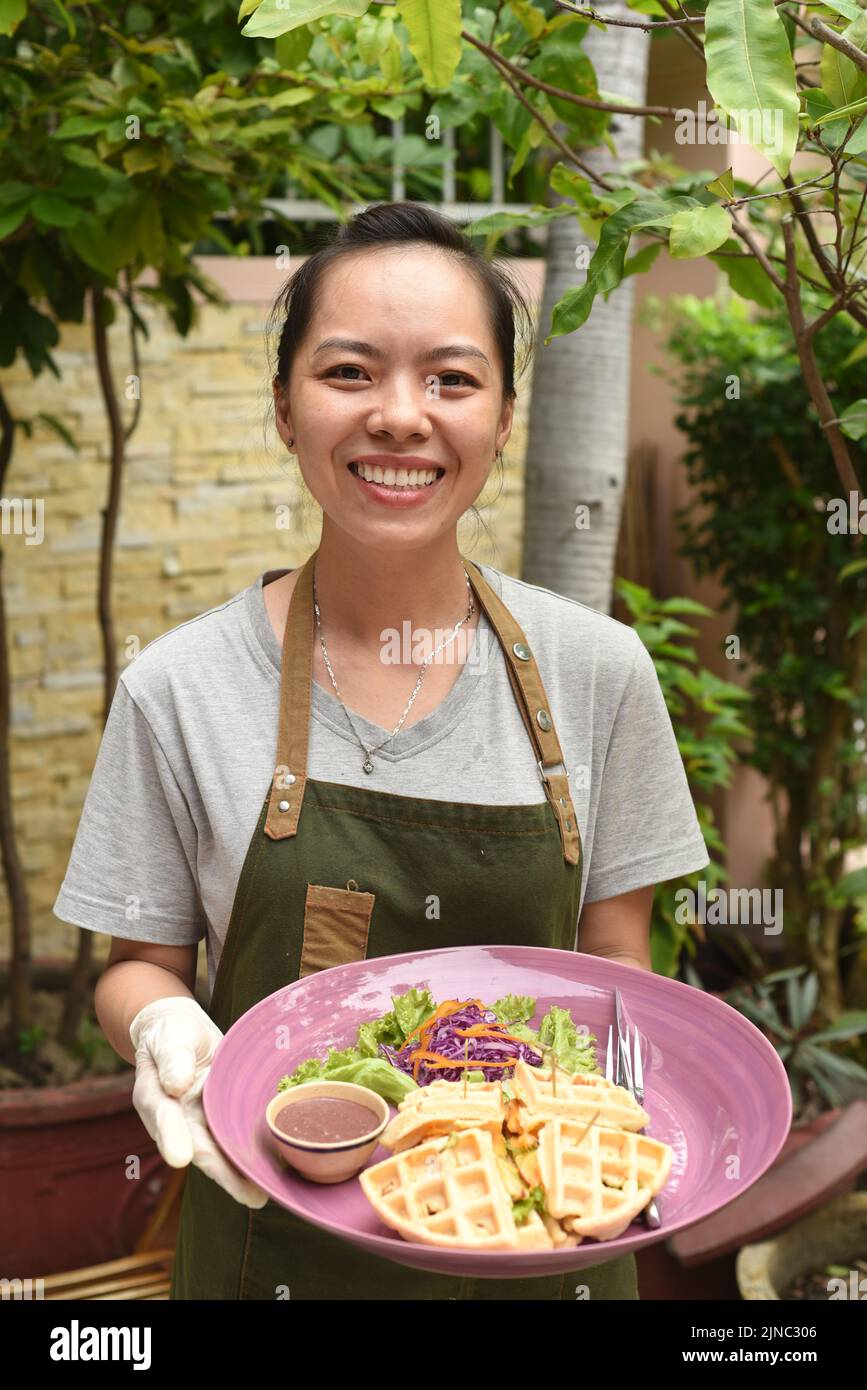 Cameriera vietnamita che serve waffle belgi con green e pollo nella caffetteria all'aperto Foto Stock