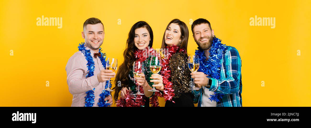 Due ragazzi e due ragazze nel tinsel di Capodanno e bicchieri di vino o champagne su sfondo giallo. Banner di Capodanno con un gruppo di studenti 20-25s Foto Stock