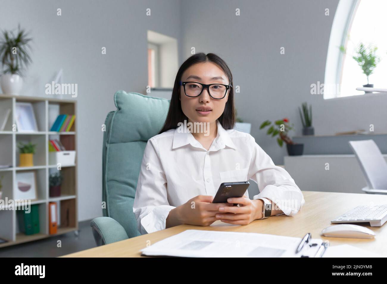 Una giovane bella donna asiatica d'affari in occhiali sta tenendo un telefono nelle sue mani, componendo un messaggio. Seduti alla scrivania in un ufficio moderno, guardando la fotocamera. Foto Stock