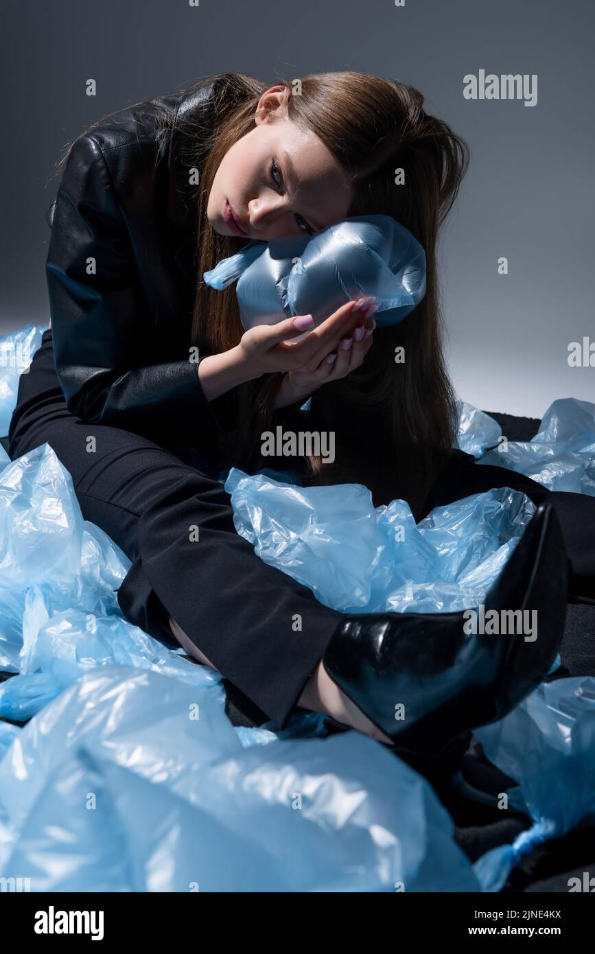 modello adolescente in elegante vestito nero che posa intorno a borse di plastica blu sul grigio Foto Stock