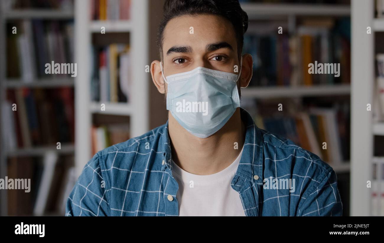 Primo piano grave giovane uomo pensivo seduto in casa da solo indossando la maschera chirurgica protettivo studente ragazzo ispanico guardando la macchina fotografica in posa in medicina Foto Stock