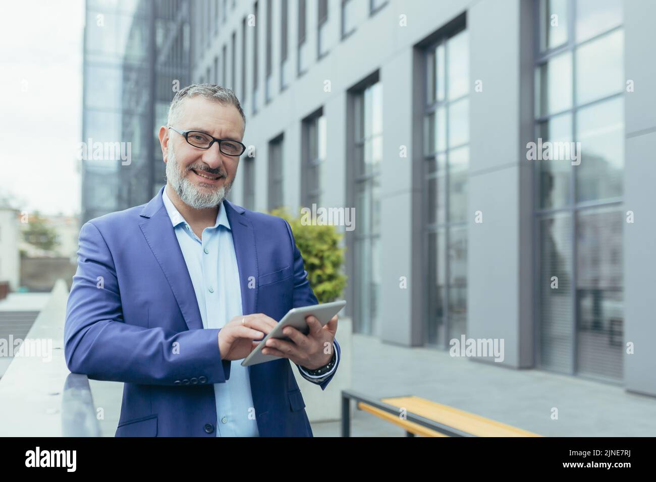 Ritratto di un uomo d'affari di successo al di fuori degli uffici con tablet in mano, investitore in occhiali e tuta aziendale sorridente e guardando la fotocamera, soddisfatto e fiducioso banchiere vicino alla banca Foto Stock
