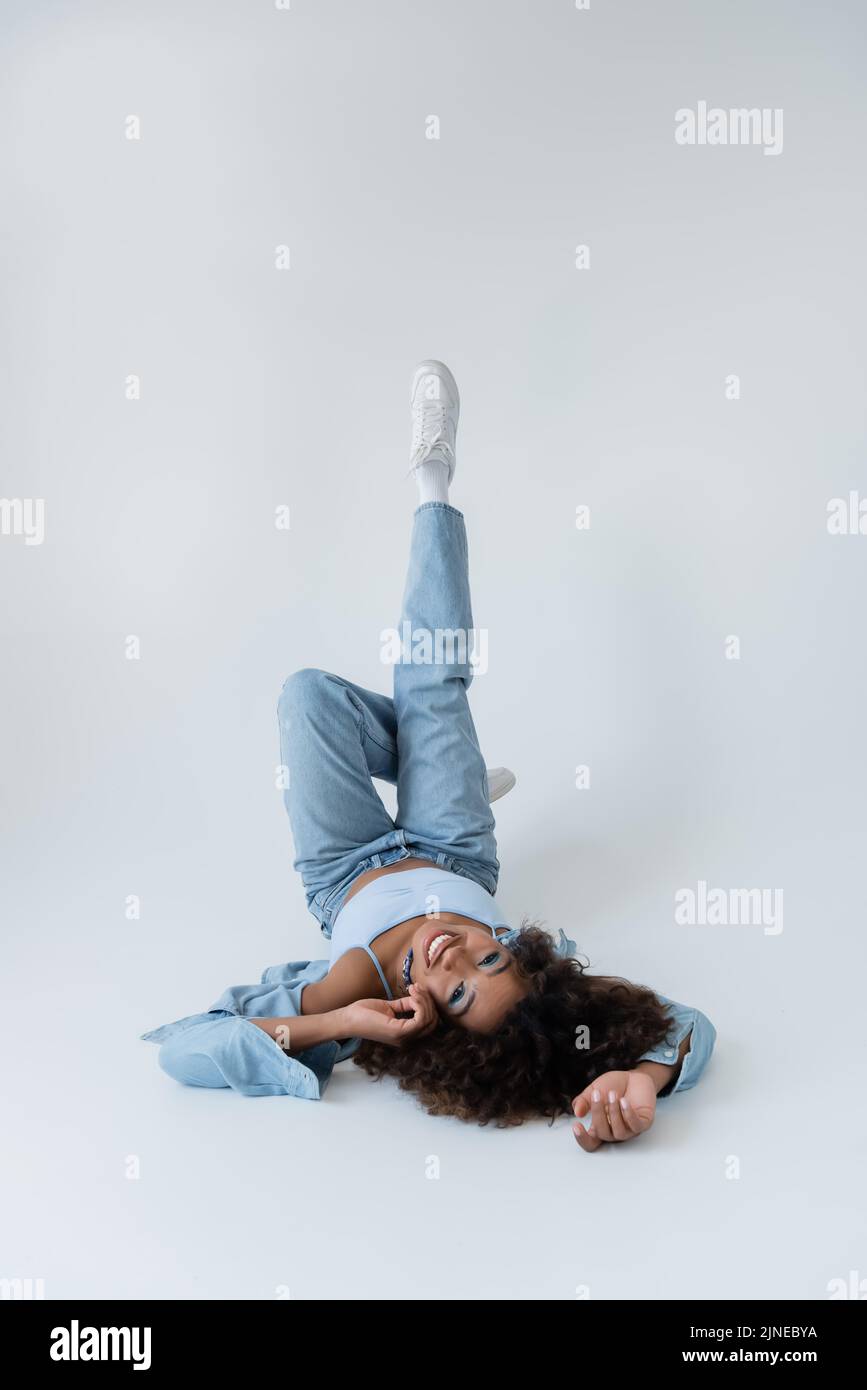 donna afroamericana sorridente sdraiata sulla schiena con gambe sollevate su sfondo grigio Foto Stock